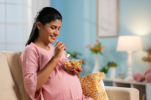 Mulher grávida sentada no sofá com um pote de frutas em sua mão. - Mustela Brazil - 1