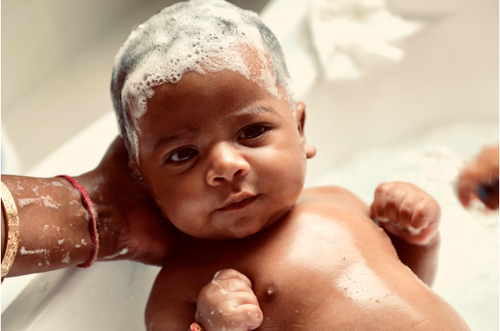 Bebê em banheira com a cabeça cheia de espuma. Suas mãozinhas estão fechadas e uma mão adulta oferece apoio para sua cabeça. - Mustela Brasil - 1