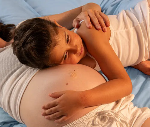 Menina deitada sobre a barriga da sua mãe grávida, abraçando-a. A mãe está com o braço gentilmente apoiado nas costas e ombros da menina. - Mustela Brasil - 1