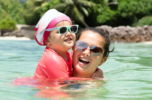 Mulher carrega e abraça menina dentro do mar. Ambas estão de óculos escuros e felizes. - Mustela Brazil - 1