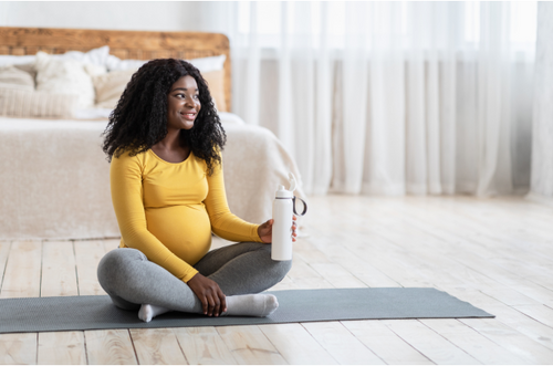 Mulher grávida sentada de pernas cruzadas sobre um tapete de exercícios, sua expressão é alegre e ela está segurando uma garrafa de água. - Mustela Brazil - 1