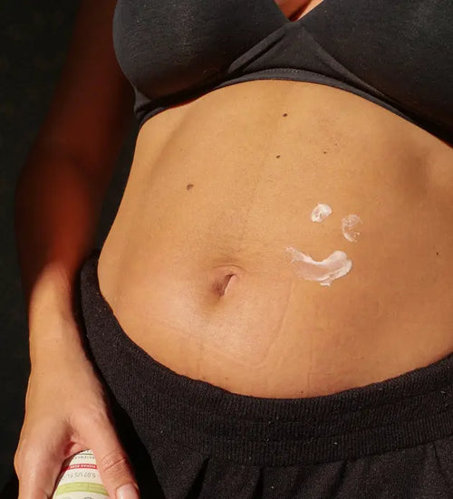 Grávida de sutiã e calça com um sorriso desenhado na barriga com creme hidratante. - Mustela Brasil - 1
