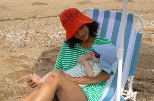 Bebê de chapéu e óculos de sol no colo de sua mãe que está sentada em uma cadeira na praia. - Mustela Brasil - 1