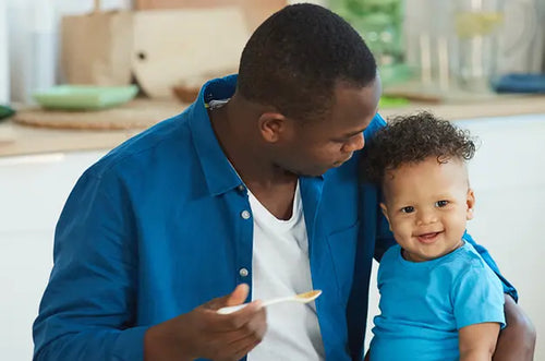 Pai segurando uma colher com alimento e oferecendo ao bebê sorridente. - Mustela Brazil - 1