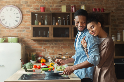 Mulher abraça carinhosamente um homem pelas costas enquanto o mesmo sorri e prepara uma refeição. - Mustela Brasil - 1