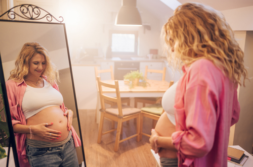 Mulher grávida acariciando sua barriga, também aparece sua imagem refletida em um espelho. - Mustela Brasil - 1
