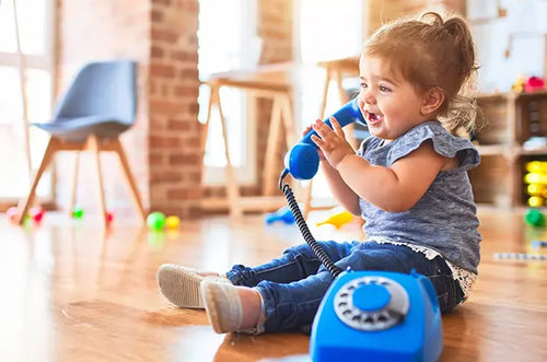 Bebê menina está sentada no chão brincando de falar no telefone. - Mustela Brasil - 1