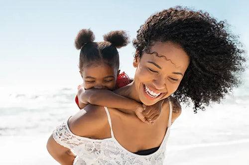Mãe carrega criança em suas costas, ambas estão sorridentes na faixa de areia com o mar ao fundo. - Mustela Brasil - 1