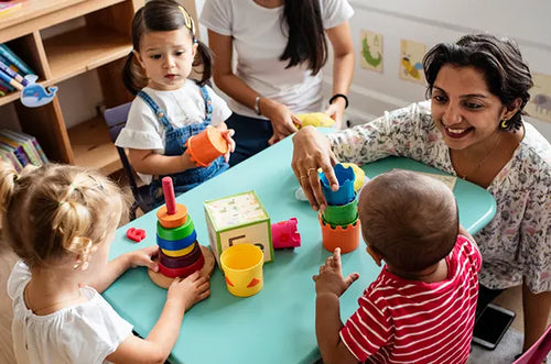 3 crianças brincam com a professora com brinquedos de madeira que estão sobre uma mesa. - Mustela Brasil - 1
