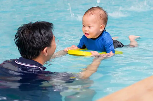 Pai auxilia bebê que está em uma pequena prancha dentro da piscina em uma aula de natação. - Mustela Brasil - 1