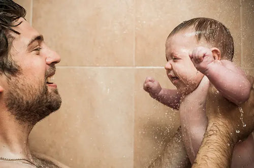 Pai segurando bebê em baixo da água do chuveiro. - Mustela Brasil - 1