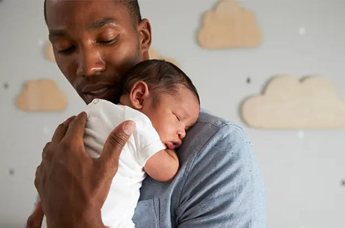 Pai segura cuidadosamente bebê recém-nascido com a mão nas costas dele. - Mustela Brasil - 1