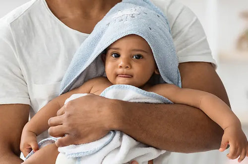 Pessoa segura bebê enrolado em toalha de banho no colo. - Mustela Brasil - 1