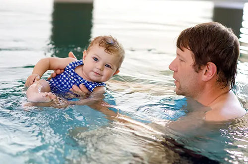 Pai segura bebê de maiô dentro da piscina em aula de natação. - Mustela Brasil - 1