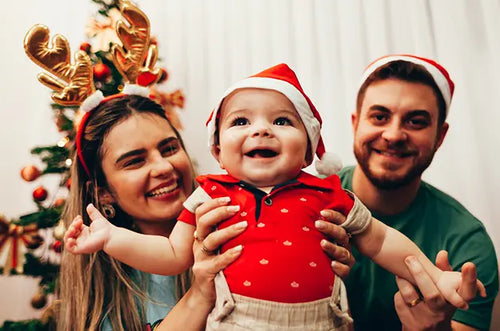 Mãe, bebê e pai sorridentes com gorro de papai Noel e ao fundo uma árvore de Natal. - Mustela Brasil - 1