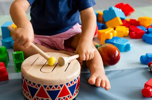 Bebê sentado no chão batucando em um tambor de madeira. - Mustela Brasil - 1