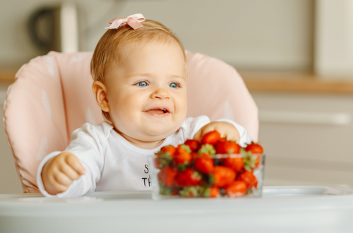 Bebê sorridente em cadeirão de alimentação com a mão em um pote cheio de morangos. - Mustela Brasil - 1