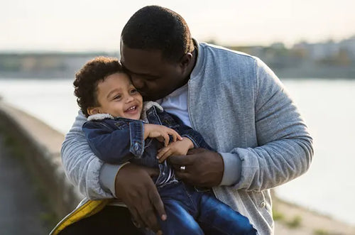 Pai abraça e beija carinhosamente filho que está em seu colo. Ambos estão com blusa de frio. - Mustela Brasil - 1