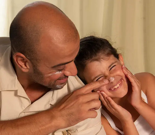 Pai brincalhão passando hidratante na ponta do nariz da sua filha que sorri com a diversão. - Mustela Brasil - 1