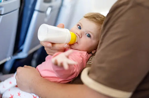 Pai em avião segura bebê no colo enquanto dá mamadeira pra ela. - Mustela Brasil - 1